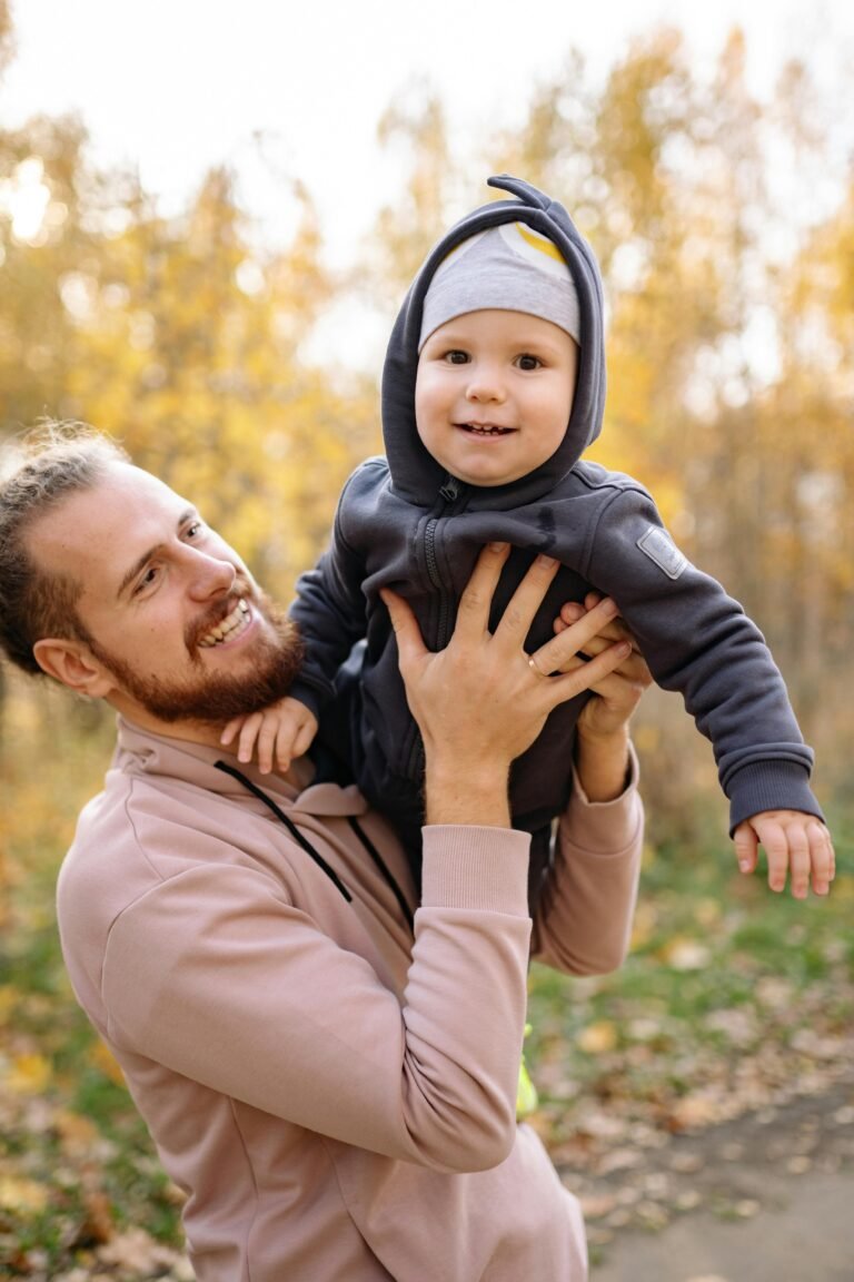 A joyful father holds his smiling baby during a sunny fall day in the park.